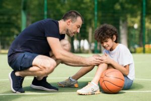 Side view portrait of focused adult man coach helping boy with knee trauma after playing basketball on the court, sad player feeling pain, touching leg. Activity And Sports Injury Concept.
